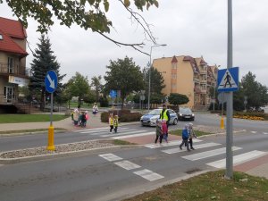 na zdjęciu jest skrzyżowanie o ruchu okrężnym, po przejściu dla pieszych przechodzą dzieci pod nadzorem umundurowanego policjanta. W tel stoi policyjny radiowóz
