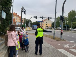 na zdjęciu, które zostało zrobione na skrzyżowaniu ulic Warszawskiej i Bohaterów Westerplatte w Giżycku widoczny jest po prawej stronie umundurowany policjant, a po lewej stronie grupa przedszkolaków. Policjantka rozmawia z dziećmi o sygnalizacji świetlnej znajdującej się na skrzyżowaniu